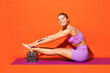 Full body young fitness trainer woman sportsman wearing top shorts purple clothes train in home gym sit do stretch hand leg exercise isolated on plain orange background. Workout sport fit abs concept.