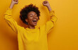 Photo of african american women does lucky dance, raises hands up in hooray, feels like champion after getting triumph, gazes happily somewhere, has fun, feels rthythm of music, isolated on yellow wal
