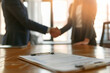 © AnastasiiaRozhmanova - sophisticated photo of a businesswoman and businessman shaking hands in the background, with a contract laying prominently in the foreground, soft white light,