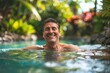 Adult man with a broad grin, enjoying a refreshing swim in crystal-clear waters, surrounded by lush greenery and vibrant tropical blooms, the epitome of relaxation and happiness