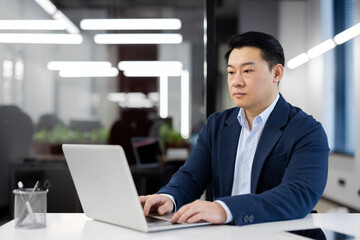 Wall Mural - A serious young Asian man in a business suit is sitting in a modern office at a desk and working on a laptop.