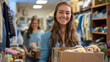 cheerful young woman is holding a box of donated items in a thrift store setting, with another person visible in the background.