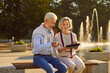 © Studio Romantic - Portrait of happy smiling senior couple sitting outdoors on bench in city park using digital tablet for social media, searching information, watching funny movie or having online video call.