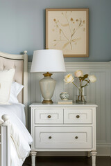 A white nightstand with two drawers and an elegant lamp on top, the wall is painted in light blue color, there's a framed art print hanging above it, the bed has beige headboard and crisp white linens