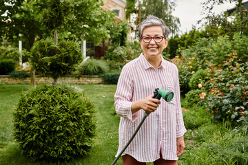 attractive joyous mature woman in casual attire watering her vibrant plants and smiling at camera