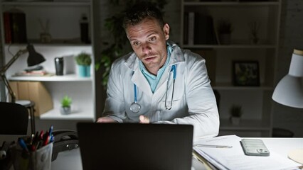 Wall Mural - Young hispanic male doctor in medical coat engaged in a video call at night inside an office, showing focused and smiling expressions.