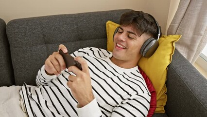 Poster - Young, confident hispanic man winning game on his gadget while lying on the comfortable sofa, exuding a sense of triumph, joy piercing his handsome portrait as he celebrates at home.