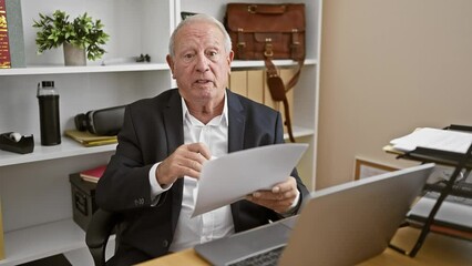 Canvas Print - Serious, mature gentleman hard at work, elegant white-haired senior man, a seasoned business professional, engrossed in documents while speaking earnestly at the office.