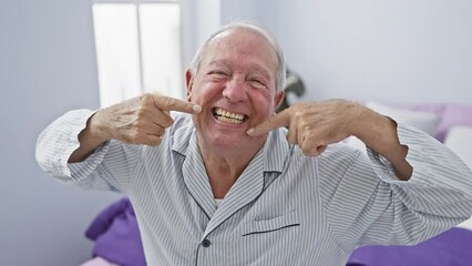 Canvas Print - Cheerful senior man in pyjamas highlights dental health, sitting on bed, pointing to perfect white teeth and showing a happy smile in a cozy bedroom.