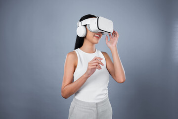 Smart female standing with gray background wearing VR headset connecting metaverse, futuristic cyberspace community technology. Elegant woman excited seeing generated virtual scenery. Hallucination.