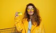 A trendy woman in a yellow coat and matching sunglasses holds a credit card, her stylish demeanor poised for a shopping spree against a vibrant yellow backdrop.