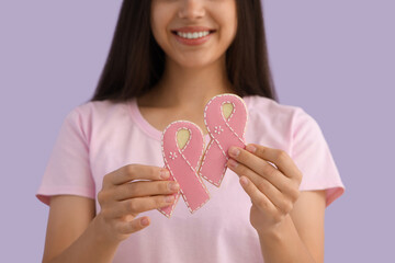 Poster - Young woman with cookies in shape of pink ribbons on lilac background, closeup. Breast cancer awareness concept