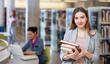 © JackF - Young woman successful student standing with books in library, other students choosing books and working on background behind
