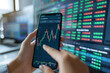 Close-up of hands using a smartphone to monitor and analyze live stock market data against a screen with financial information.
