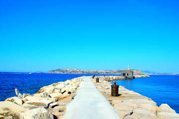 Wall Mural - Pristine view from Isola di Procida at a pier on the Tyrrhenian Sea surrounded by rocks, with a masonry building and a green lighthouse, opening to a vision of the Gulf of Naples crossed by a boat