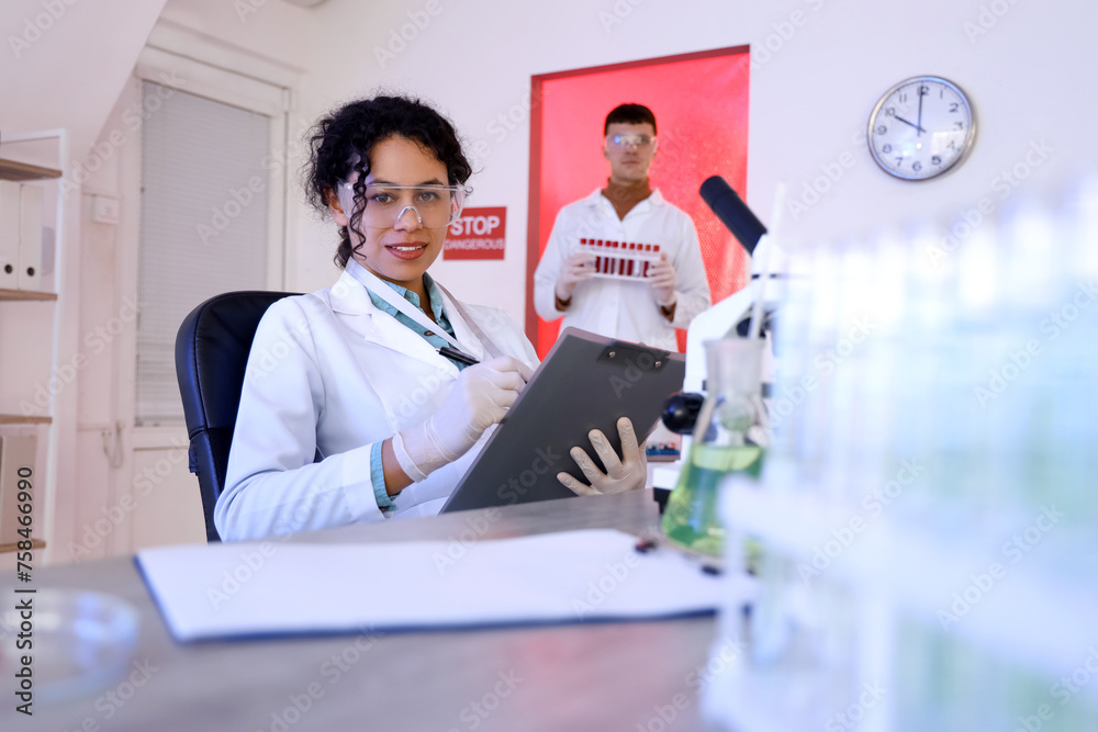 Female African-American chemist with clipboard working at table in laboratory