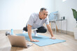 © Queenmoonlite Studio - Senior Asian man with sportswear, training and stretching doing exercise at home looking at sport video on laptop computer