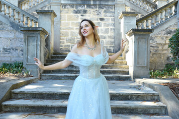 Wall Mural - Close up  portrait of beautiful female model wearing blue fantasy ballgown, like a fairytale elf princess.  Elegant pose, gestural hands reaching out, on the terrace of a romantic castle staircase 