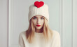 A young woman with bright red lips wearing a white beanie with a red heart on it poses in front of a light background