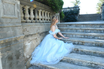 Poster - full length portrait of beautiful female model wearing blue fantasy ballgown, like a fairytale elf princess. Seated pose, sitting on staircase of a romantic castle balcony location.