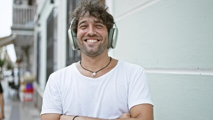 Wall Mural - Smiling hispanic man with curly hair wearing headphones and a white t-shirt standing on an urban street.