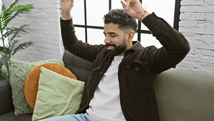 Sticker - A smiling young man with a beard sits on the couch in a cozy living room, expressing positivity and comfort.