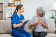 © NanSan - Caucasian teenager female doctor consults with and Asian elderly patient on medical history, discussing headaches, chest pain, coughing, sneezing, stomach pain, and neck pain while seated on a sofa.