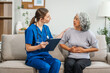 © NanSan - Caucasian teenager female doctor consults with and Asian elderly patient on medical history, discussing headaches, chest pain, coughing, sneezing, stomach pain, and neck pain while seated on a sofa.