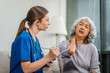 © NanSan - Caucasian teenager female doctor consults with and Asian elderly patient on medical history, discussing headaches, chest pain, coughing, sneezing, stomach pain, and neck pain while seated on a sofa.