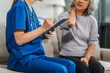 © NanSan - Caucasian teenager female doctor consults with and Asian elderly patient on medical history, discussing headaches, chest pain, coughing, sneezing, stomach pain, and neck pain while seated on a sofa.