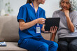© NanSan - Caucasian female doctor conducts a check-up using a tablet device for an elderly Asian patient seated together on the sofa.