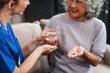 © NanSan - Caucasian female doctor provides a glass of water and medication pills to an elderly Asian patient while they are seated together on the sofa.