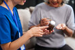 © NanSan - Caucasian female doctor provides a glass of water and medication pills to an elderly Asian patient while they are seated together on the sofa.