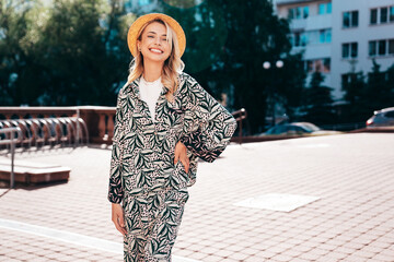 Young beautiful smiling blond hipster woman in trendy summer hippie costume clothes. Carefree female posing in the street at sunny day. Positive model outdoors at sunset. Cheerful and happy in hat