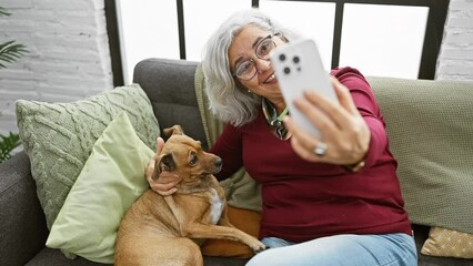 Wall Mural - A smiling elderly woman takes a selfie with her dog on a cozy sofa inside a living room.