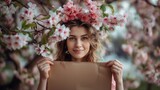 Fototapeta  - Sceneria rozkwitu wiosny. Młoda kobieta stojąca wiosną trzyma pustą kartkę przed sobą, patrząc na aparat. Jest uśmiechnięta i skupiona na treści kartki. 