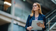 © VK Studio - Confident businesswoman with glasses holding a tablet stands in a modern corporate environment.