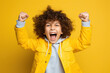 A jubilant kid model celebrating a victory, against a solid wall of yellow background, raising hands in triumph and exuding joy and happiness.