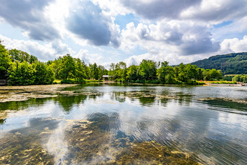 Wall Mural - Echternach lake in panoramic nature landscape, algae and reflections on water surface, green leafy trees against blue sky with white clouds in background, sunny summer day in Luxembourg