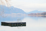 Fototapeta Lawenda - Spokój nad górskim jeziorem zimą. Drewniana platforma ze srebrną drabinką dla pływaków i plażowiczów. W tle zamglone góry.
