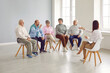 © Studio Romantic - Psychological support group for elderly and lonely people in community centre. Mature men and women talk to young female psychologist during group therapy session. Elderly people sit in row on chairs.
