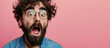 A man with a beard and glasses is looking surprised. He is wearing a blue shirt and jeans. close-up of a young man with a beard, looking surprised with his mouth wide open against a pink background