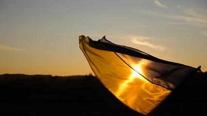 Male hand of young soldier holding blue-yellow symbol of Ukraine illuminated by sun. Ukrainian flag waving at wind outdoor at sunset. Peace and support against russian aggression. Invasion resistance