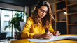 © Studio Nova - Woman with curly hair and glasses is writing in a notebook at a home office