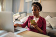 © Davor - Woman using smartphone and laptop on living room couch
