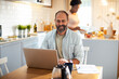 © Davor - Man smiling while working on laptop in kitchen with woman in background