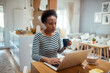 © Davor - Black woman enjoying breakfast while working on laptop in a sunny kitchen
