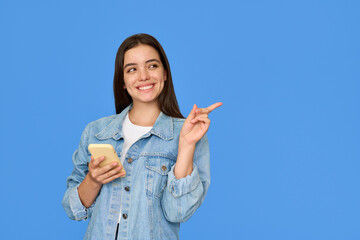 Happy pretty Latin gen z teen girl pointing aside with smartphone, smiling Hispanic customer using mobile cell phone advertising shopping sale presenting app promo on blue background.