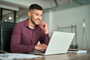 Happy busy Latin business man employee or manager working on digital technology project sitting at desk. Young smiling professional businessman entrepreneur using computer laptop in office.