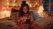 Glad Cheerful Girl Using Smartphone While Resting In Bedroom. Warm Colors, Cozy Home Mood. Wearing Pajamas.